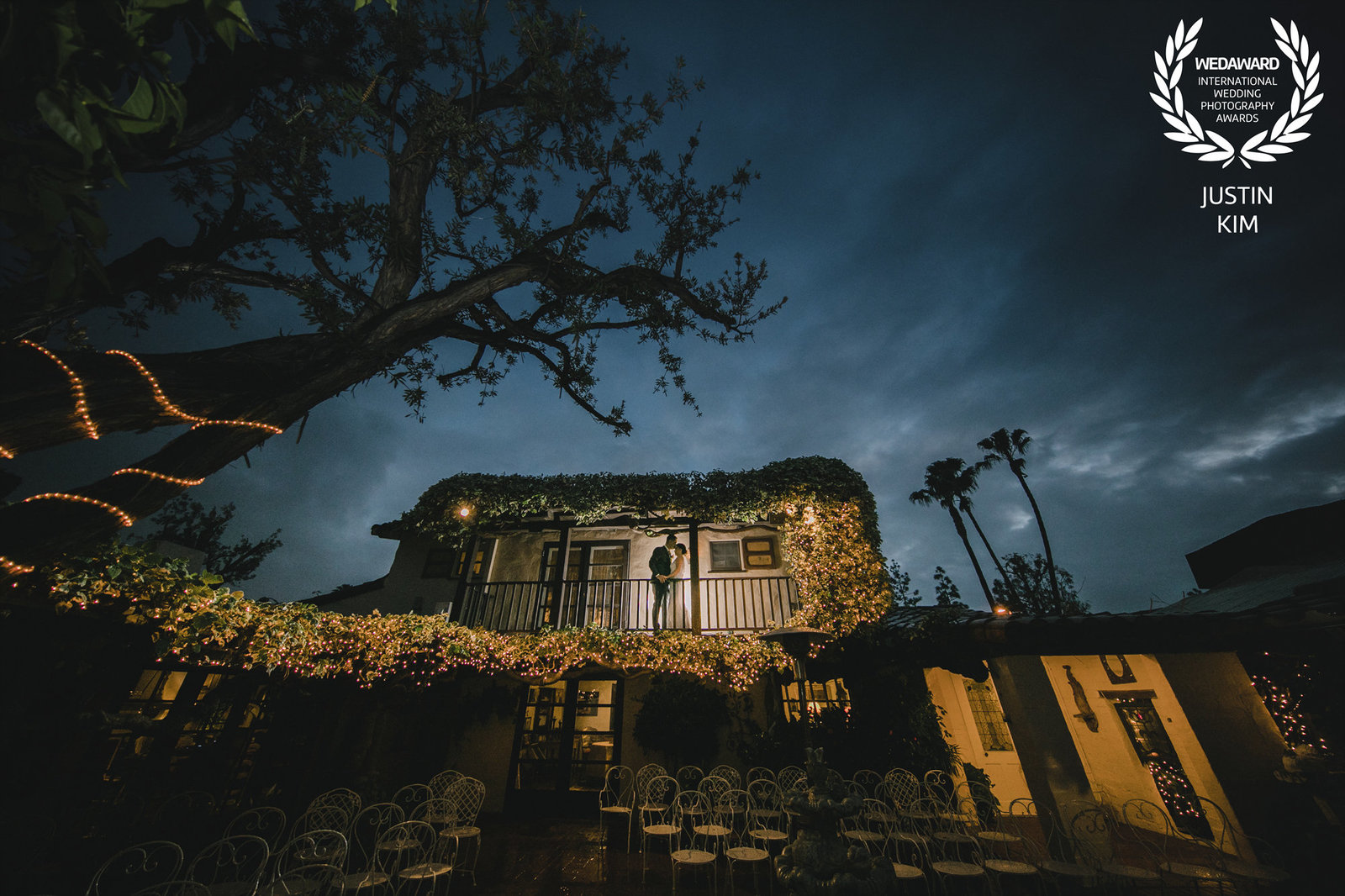 A newlywed couple sharing a quiet moment on a lit, ivy-covered balcony at their evening wedding reception